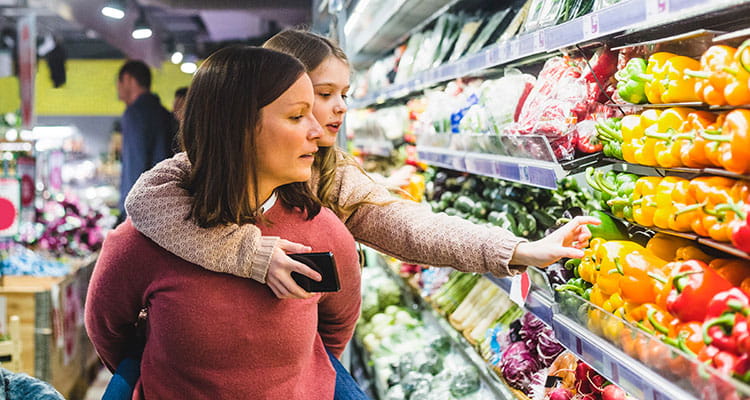 Daughter choosing bell peppers while being piggybacked by mother in grocery store.