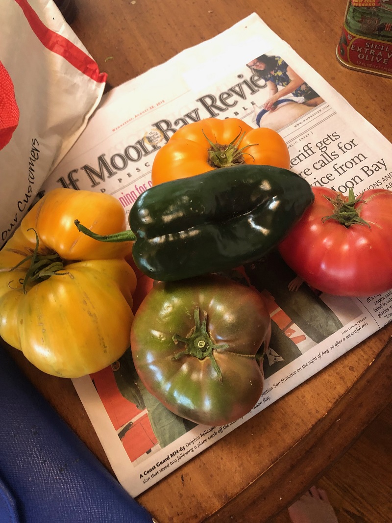 A pepper and a variety of large heirloom tomatoes are sitting on top of the Half Moon Bay Review newspaper.