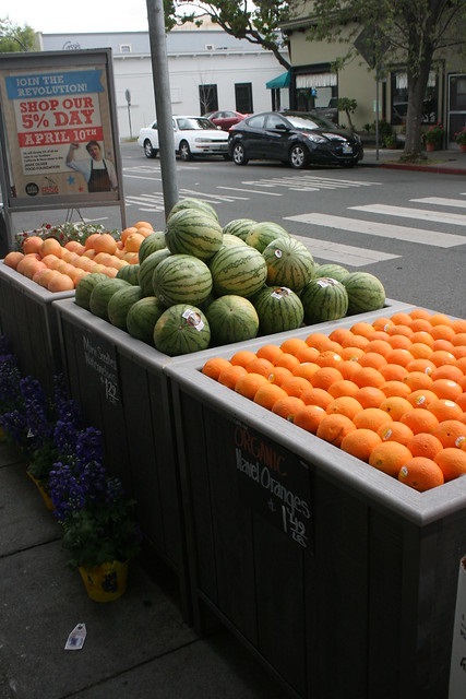 An outdoor market with containers of oranges and watermelon