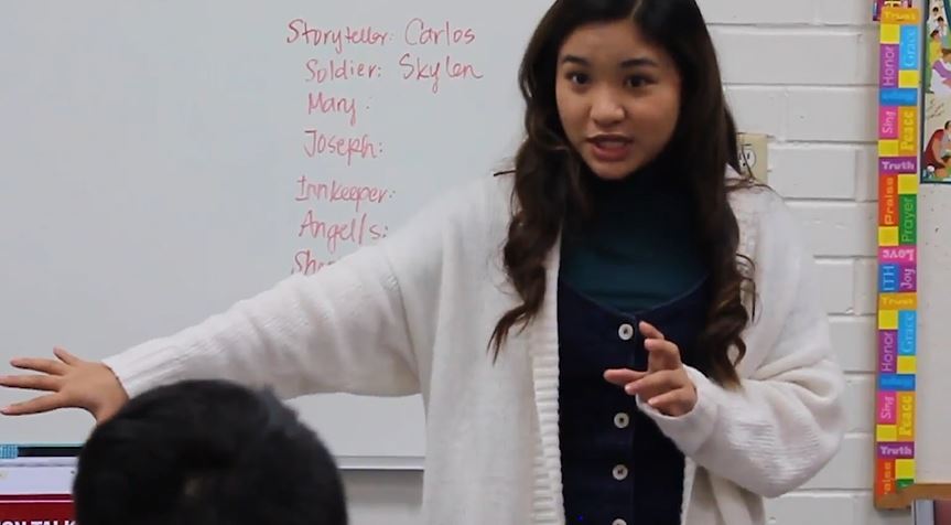 A young adult woman is speaking at the head of a classroom.